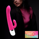 A person wearing a pink top and black coat gazes off to the side, partially shadowed. Beside them is the VeDO Wink Rabbit Vibe in Hot Pink, offering twelve gratifying modes. The bottom right corner showcases the VeDO logo accompanied by the text "let your body vibe" on a vibrant background.